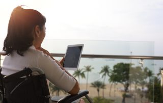 Handicapped person or patient sit on wheelchair using tablet at balcony of hotel at beachside