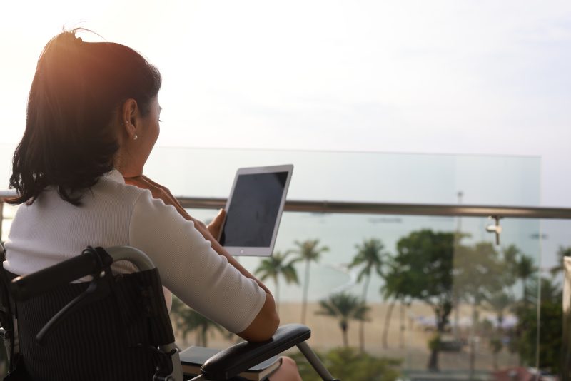 Handicapped person or patient sit on wheelchair using tablet at balcony of hotel at beachside